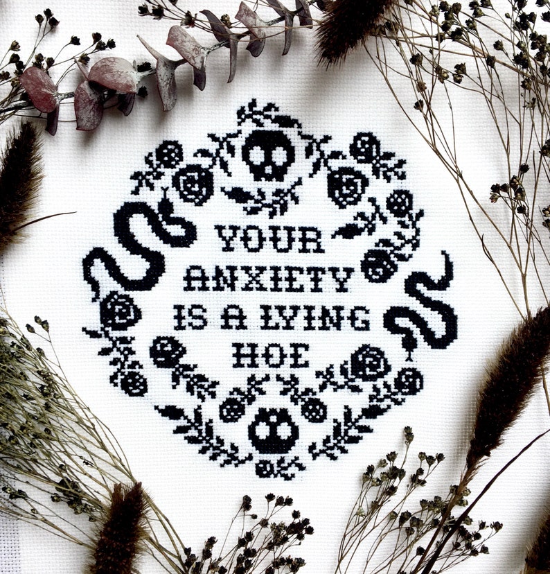 Może przedstawiać: Czarno-białe haftowane krzyżykiem z tekstem "Your anxiety is a lying hoe" otoczone wzorem kwiatowym z czaszkami i wężami.
