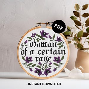 May include: A cross-stitch embroidery hoop featuring the phrase "woman of a certain rage" encircled by purple flowers and green leaves. The design is on white fabric, set in a wooden hoop. A PDF icon is in the upper right corner.