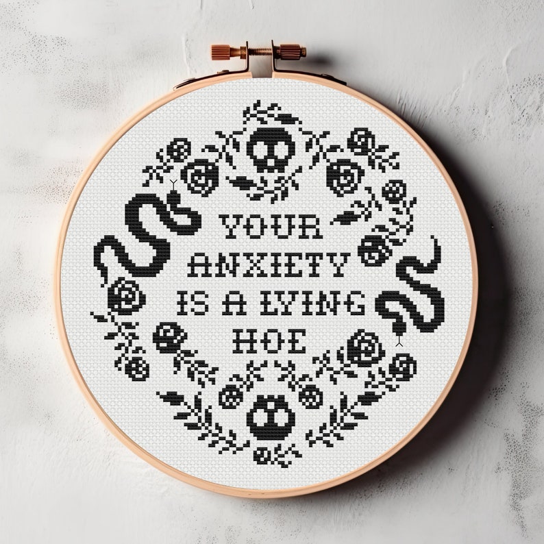 Może przedstawiać: Czarno-biały tamborek z haftem przedstawiającym napis "Your anxiety is a lying hoe" otoczony kwiatowym wzorem z czaszkami i wężami.