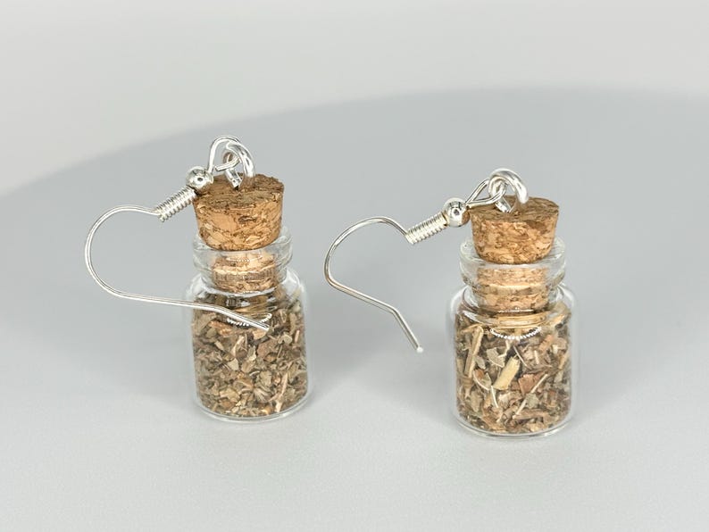 Puede incluir: Un par de pendientes de plata con peque&ntilde;as botellas de vidrio llenas de hierbas secas y tapas de corcho.