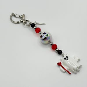 May include: A white ghost charm with a red pitchfork, a white bead with a black and blue face, and red and black beads on a silver chain with a silver clasp.
