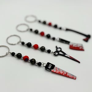 May include: Four keychains with black and red beads and different weapon charms. The charms include a saw, a pair of scissors, a knife, and an axe. Each charm has red paint splatters on it.