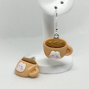 May include: A pair of dangle earrings featuring a brown coffee cup with a white face and a brown swirl on top. The earrings are made of polymer clay and have silver hooks.