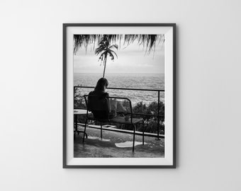 May include: Black and white photograph of a person sitting on a bench overlooking the ocean. The person is facing away from the camera and is looking out at the water. A palm tree is visible in the background.