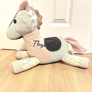 Könnte beinhalten: Ein Plüschpferd aus verschiedenen Stoffstücken in den Farben Hellrosa, Salbeigrün und Creme mit Tiermotiven. An der Seite des Pferdes ist der Name Floyd in schwarzem Garn gestickt und es trägt einen Sattel aus dunkelgrauem Stoff.