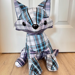 Könnte beinhalten: Ein Stofftier in Form eines Fuchses mit einem karierten Muster in Weiß, Grau, Blau und Lila. Der Fuchs hat schwarze Knopfaugen und eine schwarze Nase. Das Spielzeug sitzt auf einem Holzboden.
