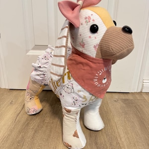 Könnte beinhalten: Ein Stoffhund aus verschiedenen Stoffstücken mit Blumen-, Bären- und Eulenmustern. Das Spielzeug trägt ein altrosa Halstuch mit Löwenmotiv, eine gerippte rosa Schleife auf dem Kopf und einen weißen Rüschenrock im Bereich der Taille.