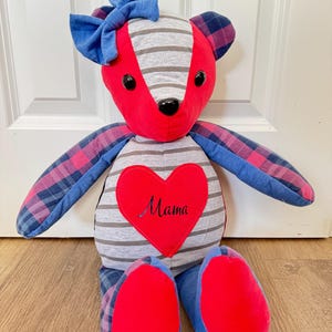Könnte beinhalten: Ein Stoffteddybär aus verschiedenen Stoffmustern, darunter grau-weiße Querstreifen, einfarbiges Rot und ein blau-rotes Karomuster. Auf dem Kopf befindet sich eine blaue Stoffschleife und auf der Brust ein rotes Herz mit dem Wort Mama.