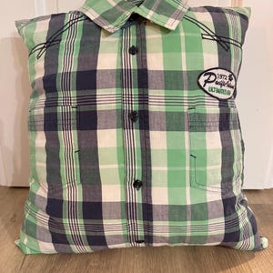 Könnte beinhalten: Ein dekoratives Kissen aus einem grün, marineblau und weiß karierten Hemd. Das Kissen hat den Kragen, die Knöpfe und einen Aufnäher mit der Aufschrift "Pacific Island Ultimate Surf". Es liegt auf einem Holzboden.