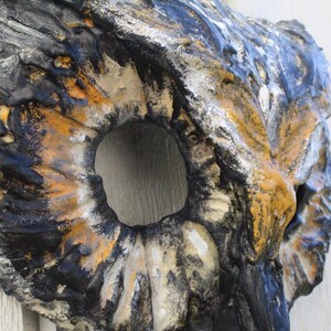 May include: A textured, abstract wall sculpture with a large central opening. The piece features a blend of colors, including black, gold, white, and blue, creating a weathered appearance. The sculpture's design suggests a natural, organic form.