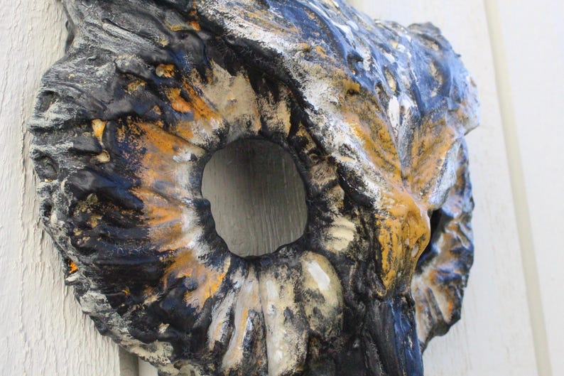 May include: A textured, abstract wall sculpture with a large central opening. The piece features a blend of colors, including black, gold, white, and blue, creating a weathered appearance. The sculpture's design suggests a natural, organic form.