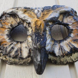 May include: A handcrafted owl mask with a detailed, textured surface. The mask features large eye openings, a prominent beak, and is painted in shades of brown, black, and white. This decorative mask is suitable for costume parties or theatrical performances.