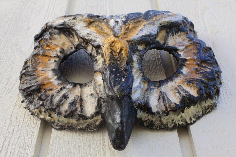 May include: A handcrafted owl mask with a detailed, textured surface. The mask features large eye openings, a prominent beak, and is painted in shades of brown, black, and white. This decorative mask is suitable for costume parties or theatrical performances.