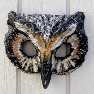 May include: A handcrafted owl mask with a textured surface. The mask features a detailed design with black, white, and brown hues. The eye openings are large and oval-shaped, and the beak is prominent. The mask is likely made of a durable material.