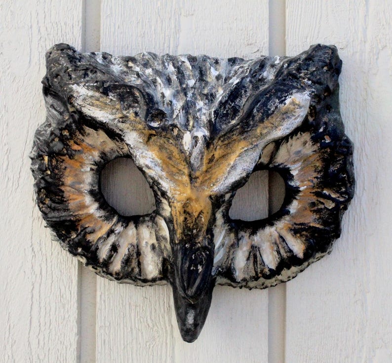 May include: A handcrafted owl mask with a textured surface. The mask features a detailed design with black, white, and brown hues. The eye openings are large and oval-shaped, and the beak is prominent. The mask is likely made of a durable material.