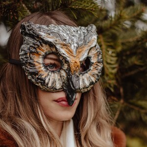 May include: An owl mask with a detailed, textured design in shades of gray, brown, and orange. The mask has large eye openings and a prominent beak. A black strap secures the mask. The mask is worn by a person with long, light brown hair.