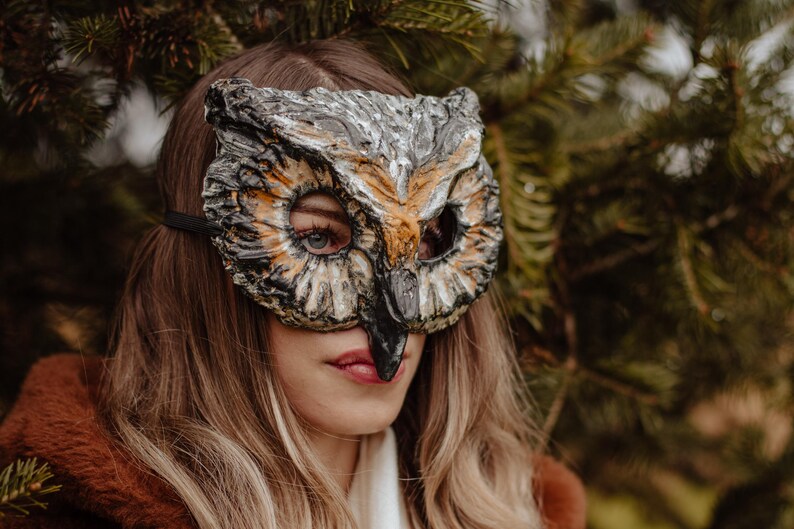 May include: An owl mask with a detailed, textured design in shades of gray, brown, and orange. The mask has large eye openings and a prominent beak. A black strap secures the mask. The mask is worn by a person with long, light brown hair.