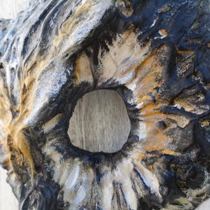 May include: A textured, abstract wall sculpture with a central circular opening. The piece features a blend of colors, including black, white, and gold, creating a weathered, organic appearance. The sculpture is mounted on a white surface.