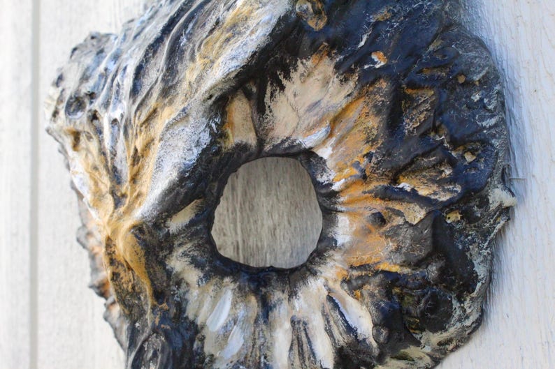 May include: A textured, abstract wall sculpture with a central circular opening. The piece features a blend of colors, including black, white, and gold, creating a weathered, organic appearance. The sculpture is mounted on a white surface.