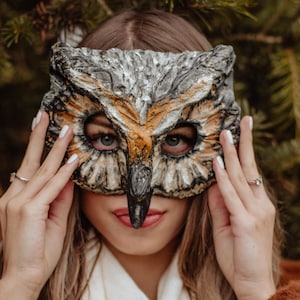 May include: An owl mask with a detailed design in shades of gray, brown, and gold. The mask covers the eyes and nose, with the wearer's eyes visible. The mask is held up by hands with white nail polish, against a blurred green background.