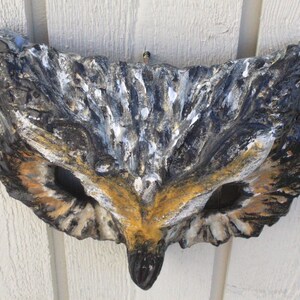 May include: A decorative owl mask made of stone, featuring a detailed design with black, white, and brown hues. The mask has large eye openings and a pointed beak, suitable for wall decor.