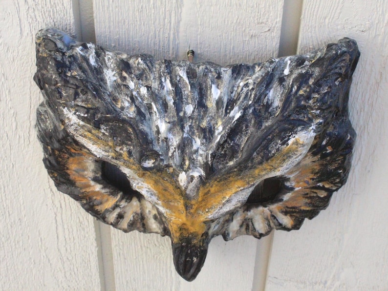 May include: A decorative owl mask made of stone, featuring a detailed design with black, white, and brown hues. The mask has large eye openings and a pointed beak, suitable for wall decor.