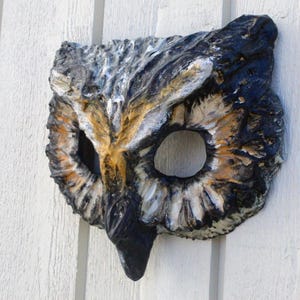 May include: A handcrafted owl mask with a textured surface. The mask features a detailed design with black, white, brown, and gold colors. The eye holes are large and round, and the beak is prominent. The mask is likely made of a durable material.
