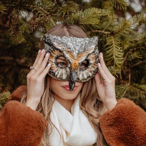 May include: An owl mask with a detailed design in shades of gray, brown, and orange. The mask is held up in front of a person's face. The person is wearing a brown coat and a white scarf, with a background of green pine needles.