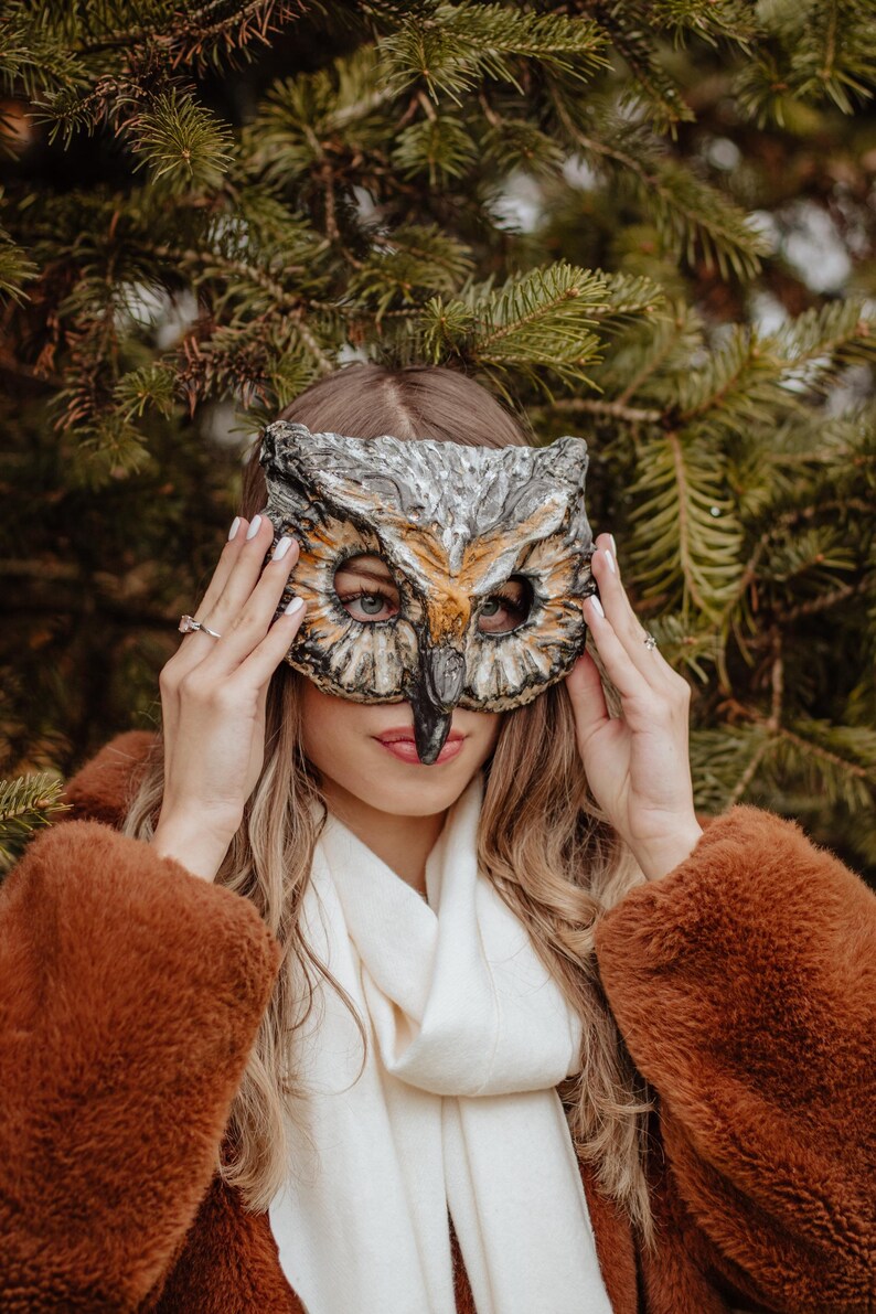 May include: An owl mask with a detailed design in shades of gray, brown, and orange. The mask is held up in front of a person's face. The person is wearing a brown coat and a white scarf, with a background of green pine needles.