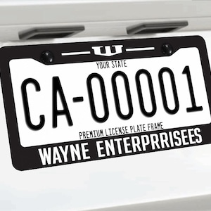 Può includere: Cornice per targa nera e bianca con il testo "CA-00001" e "WAYNE ENTERPRRISES". La cornice è montata su una superficie bianca. La parte superiore della cornice riporta la scritta "YOUR STATE".