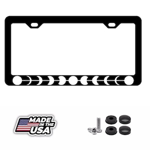 May include: A black rectangular license plate frame featuring a white graphic of the lunar cycle phases along the bottom edge. The set includes two metal screws and four black plastic screw covers, accompanied by a Made in the USA emblem.