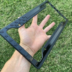 Puede incluir: Un marco negro para matrícula con un diseño de estrellas en el lado izquierdo. El marco está sostenido por una mano sobre un fondo de hierba verde. El marco tiene un acabado brillante.