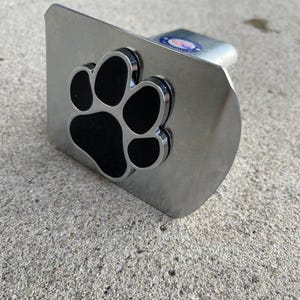 May include: A silver-colored metal trailer hitch cover featuring a black paw print design. The paw print is raised and has a polished finish. The hitch cover is rectangular with rounded edges and a small, circular emblem.
