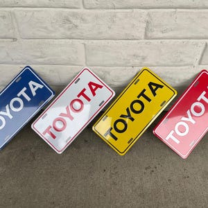 Könnte beinhalten: Vier rechteckige Nummernschilder mit dem Wort "TOYOTA" in weißen Buchstaben. Die Schilder sind blau, weiß, gelb und rot. Die Schilder sind zum Betrachter hin abgewinkelt.