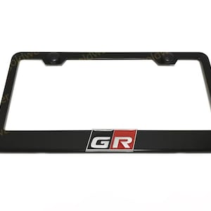 May include: A rectangular black license plate frame with rounded corners. The bottom center features a metallic emblem with the letters GR in white on a black and red background. Two black circular screw covers are positioned at the top corners.