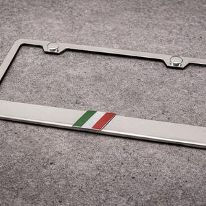 May include: A silver license plate frame with a green, white, and red stripe. The frame has a rectangular shape with rounded corners and two silver fasteners at the top.