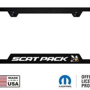 May include: A black rectangular number plate surround featuring the white text SCAT PACK and a small bee graphic on the bottom edge. The frame includes two black mounting screw covers at the top and displays the Made in the USA and Mopar logos.