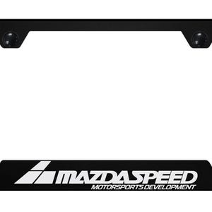 Op de afbeelding: Een rechthoekige zwarte kentekenplaathouder met de witte tekst MAZDASPEED MOTORSPORTS DEVELOPMENT op de onderrand. Het frame bevat twee ronde montagegaten in de bovenhoeken voor bevestiging aan een voertuig.