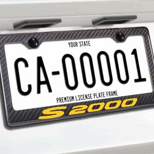 May include: A license plate with the text "CA-00001" in black on a white background, framed by a carbon fiber-style border. The bottom of the frame has the text "S 2000" in yellow. The top of the plate reads "YOUR STATE".