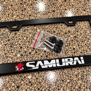 May include: A black rectangular license plate frame featuring the white text SAMURAI and a red and white graphic of a stylized oni mask. Included is a small clear plastic bag containing two silver metal screws and two black screw covers.