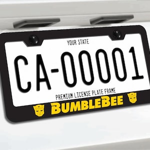 May include: A black license plate frame featuring the yellow text BUMBLEBEE and two yellow robot face icons at the bottom. The frame surrounds a white license plate with the text YOUR STATE at the top and the alphanumeric code CA-00001 in black.