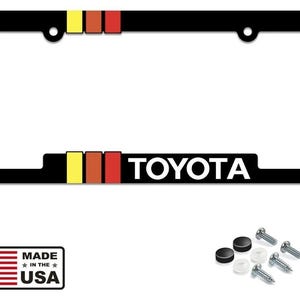 Puede incluir: Marco de matrícula negro con la palabra "TOYOTA" en blanco. El marco tiene rayas amarillas, naranjas y rojas. Incluye accesorios de montaje. Hecho en los EE. UU.