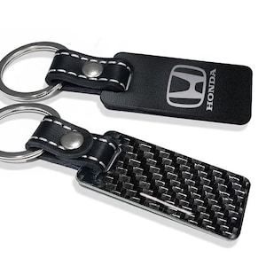 以下が含まれることがあります： シルバーの金属リングが付いた2つの長方形のキーホルダー。1つは白いステッチ、シルバーのリベット、シルバーでプリントされたHondaロゴが入った黒いレザー調のストラップ付き。もう1つは黒いカーボンファイバー柄の仕上げ。