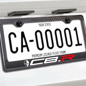 Puede incluir: Placa de matrícula con el texto "CA-00001" en negro sobre fondo blanco, enmarcada por un borde con efecto de fibra de carbono. El marco incluye texto y un logotipo.