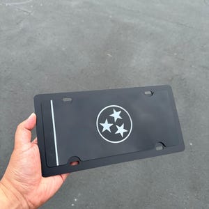 May include: Black license plate frame featuring the Tennessee state symbol, a white circle with three white stars. A vertical white stripe is on the left side. The frame has four mounting holes.