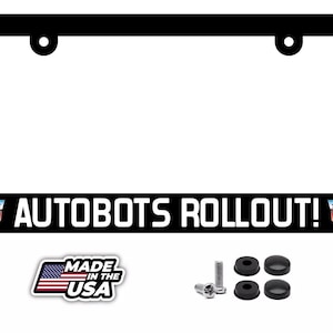 May include: A black rectangular license plate frame featuring the white text AUTOBOTS ROLLOUT! flanked by two red, white, and blue Autobot logos. Included are four black screw covers and two metal screws, with a Made in the USA flag emblem.