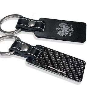 May include: Two rectangular keychains with silver metal rings. One keychain features a black surface with a white eagle emblem. The other keychain has a black and gray carbon fiber pattern. Both have black leather straps with white stitching and silver rivets.
