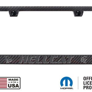 May include: A carbon fiber license plate frame with the word "HELLCAT" in silver lettering. The frame has a textured pattern and includes the Mopar logo and "OFFICIAL LICENSED PRODUCT" text. The frame is made in the USA.