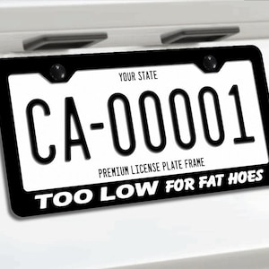 May include: A black license plate frame with white text that reads "TOO LOW FOR FAT HOES" along the bottom edge. The frame surrounds a white license plate featuring the text "YOUR STATE", "CA-00001", and "PREMIUM LICENSE PLATE FRAME".