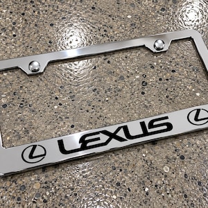 Puede incluir: Un marco rectangular para matrícula de cromo con un acabado pulido y reflectante. La palabra LEXUS está impresa en letras mayúsculas negras en la parte inferior central, flanqueada por dos logotipos circulares negros de Lexus.